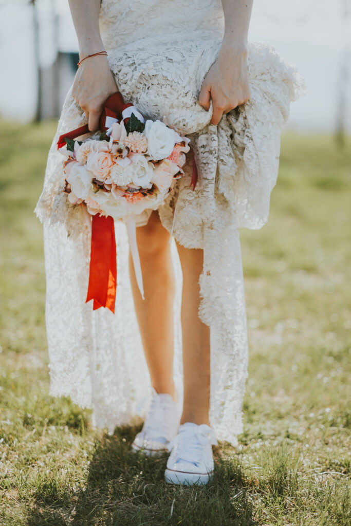 Frau im Boho-Hochzeitskleid mit Blumenstrauß in der Hand trägt niedrige weiße Sneakers