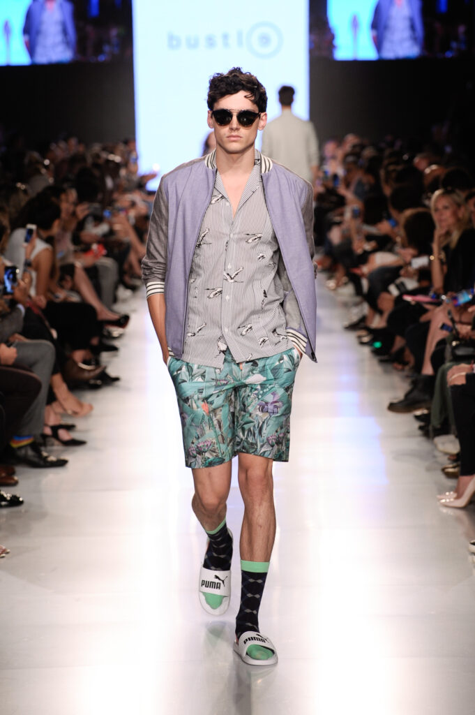 Model gekleidet im Sommer-Outfit: gemustertes Hemd und Shorts, violettfarbene Bomberjacke und weiße Puma Pantoletten kombiniert mit gemusterten Socken