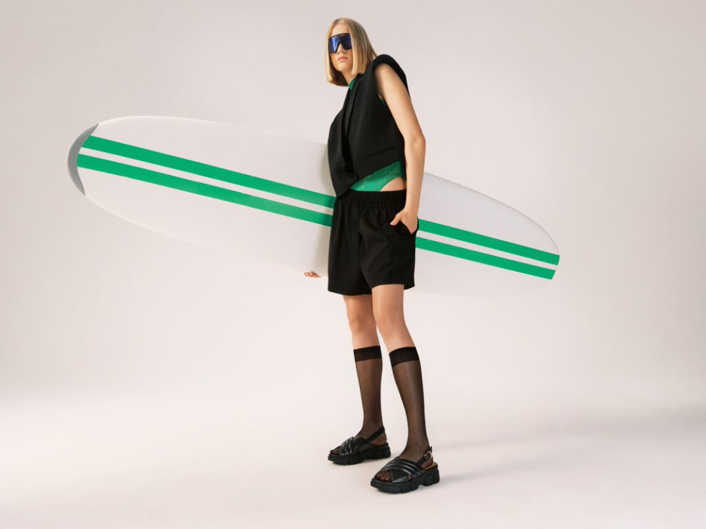 Model im schwarzen Total-Look mit schwarzen Sandalen, ein Surfbrett in der Hand - Sommer 2023 Kampagne