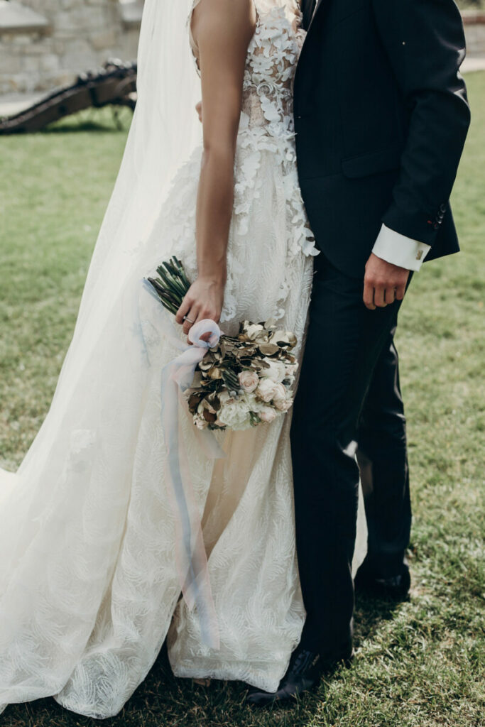 Stilvolle Braut hält einen goldenen Strauß aus Rosen mit Bändern, steht und umarmt den Bräutigam